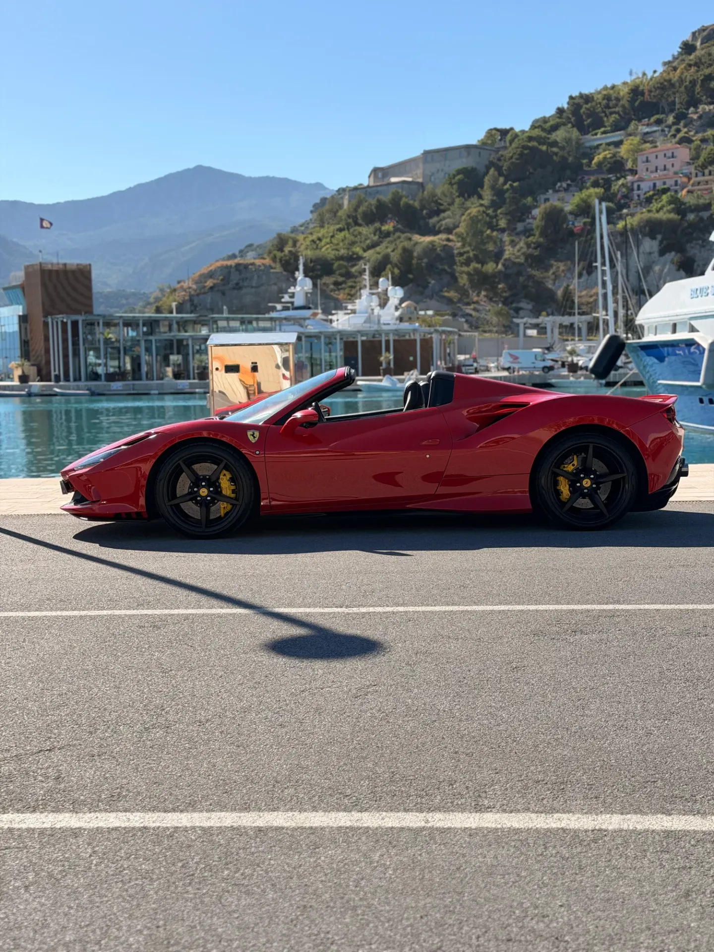 Red Ferrari F8 in side profile beside the yachts at a marina.