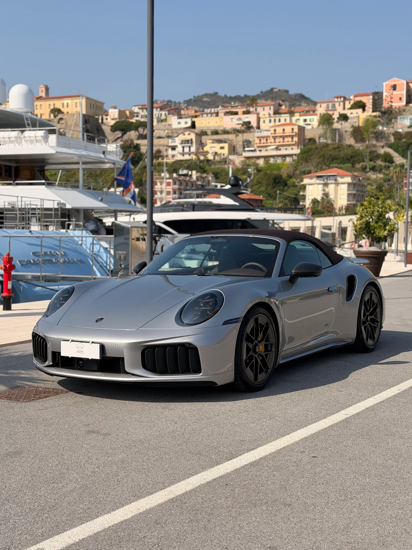 Silver Porsche 911 Turbo S with a dark roof, seen from the front three-quarter angle at a marina.